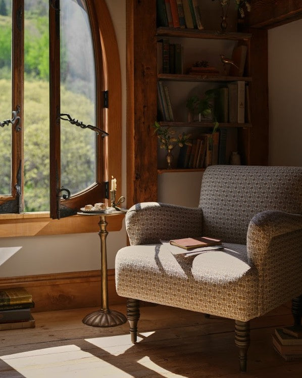 A cozy upholstered armchair in a rustic room with warm wood paneling, a window with outdoor greenery, a small side table, and a book on the chair.
