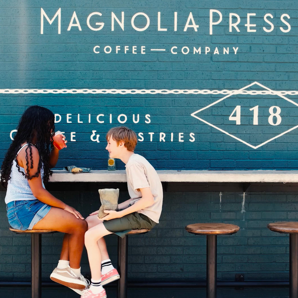 Two children sitting on stools at a cafe counter in front of a painted brick wall with large white text reading 'Magnolia Press Coffee Company' and '418'.