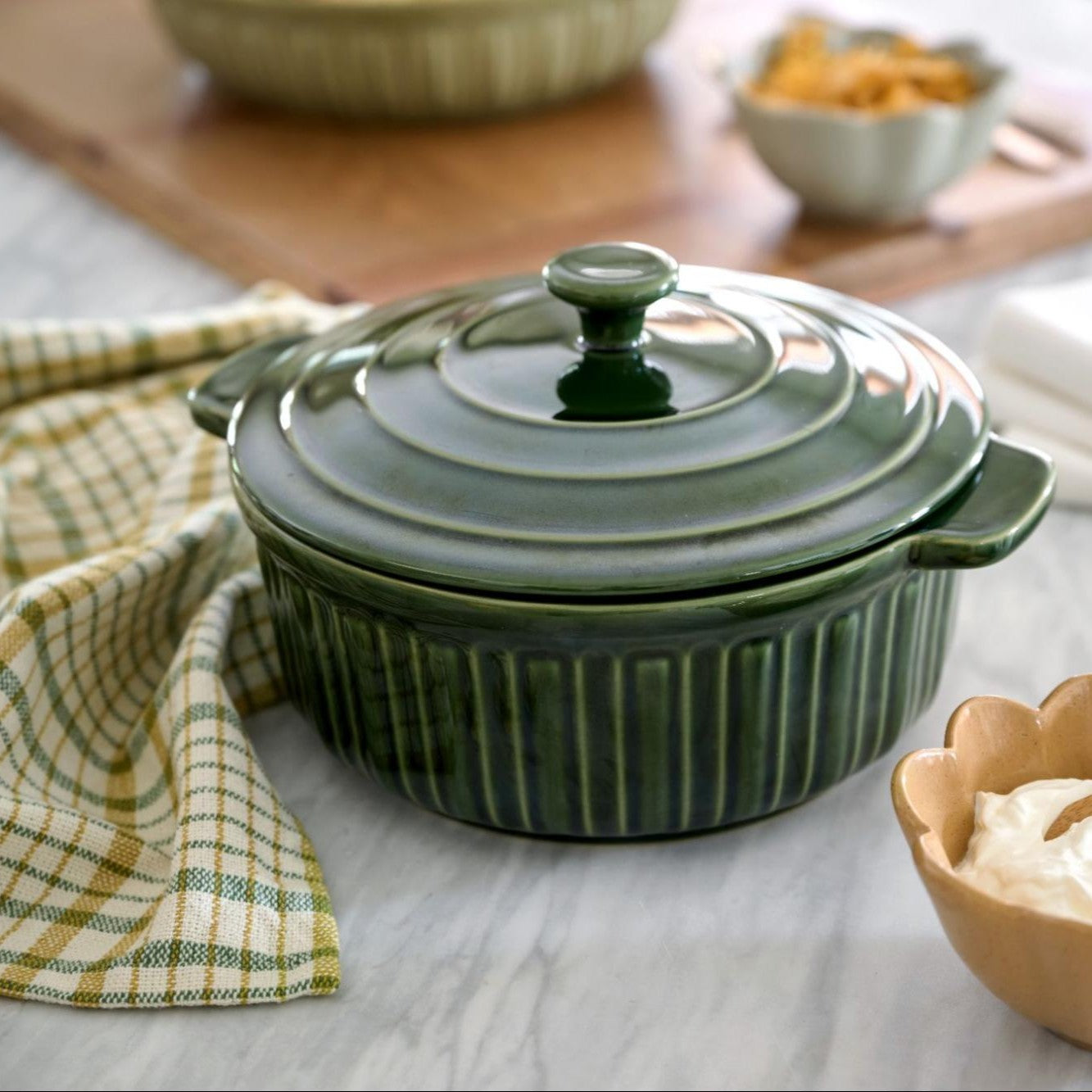 Green ceramic baking dish with lid on a wooden surface with a checkered cloth and bowls in the background.