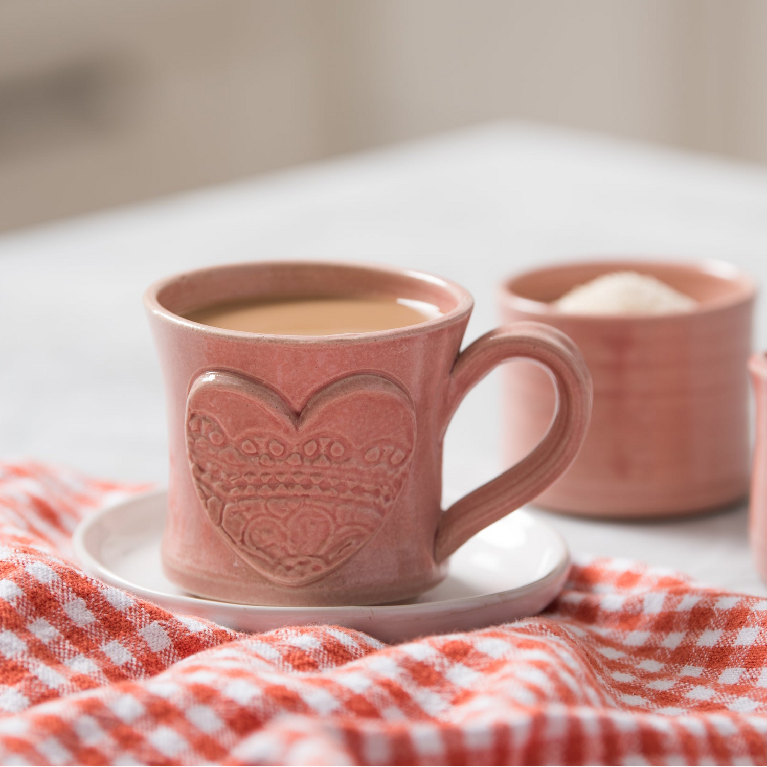 Pink mug with heart design on a white surface with a red and white checkered cloth.