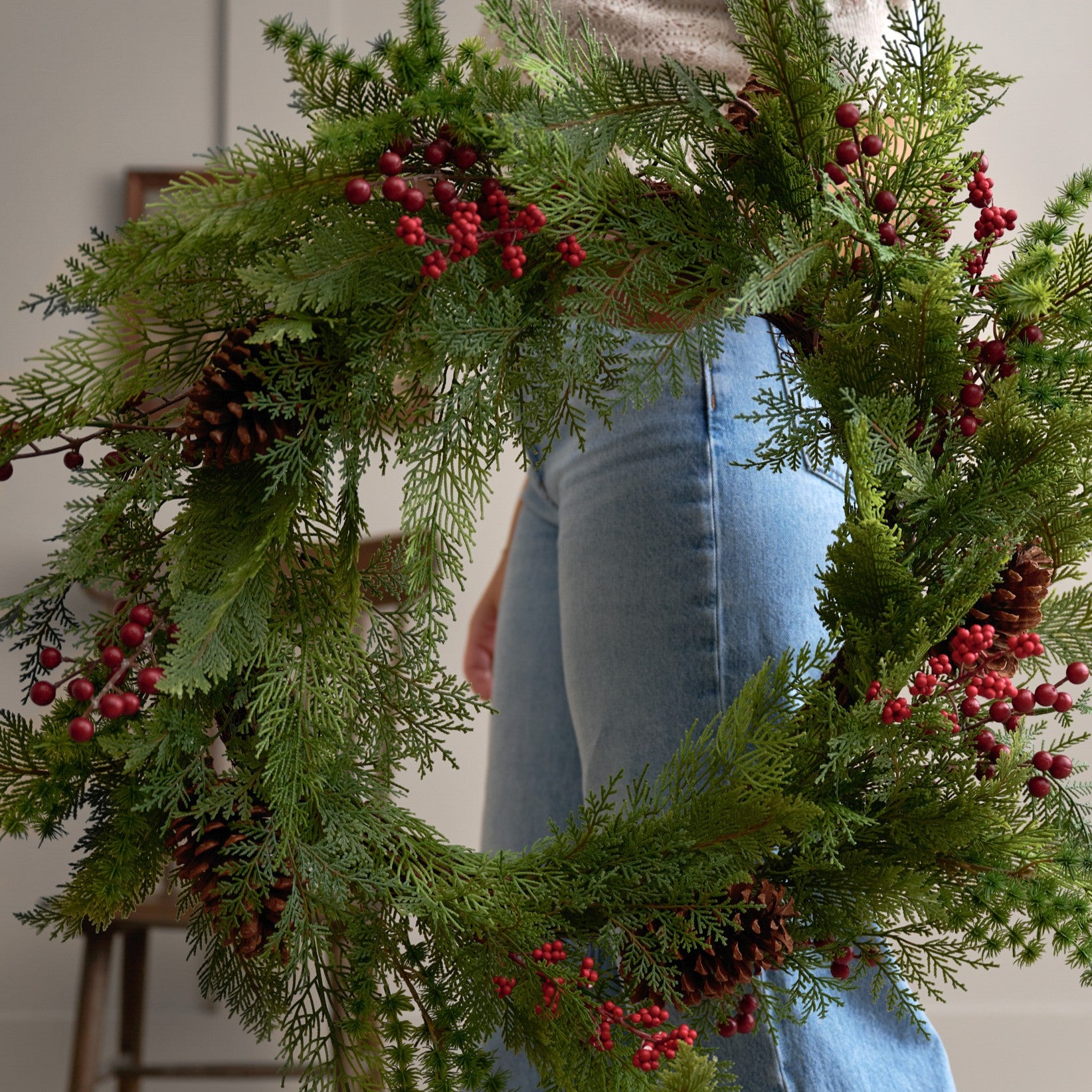 person holding a greenery wreath with red berries and pinecones