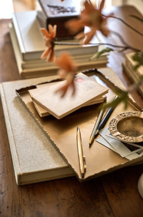 A styled flat-lay of vintage stationery on a wooden table, featuring a linen-covered notebook, a stack of old books, a gold pen, a small metal inkwell, and dried orange flowers in the foreground.
