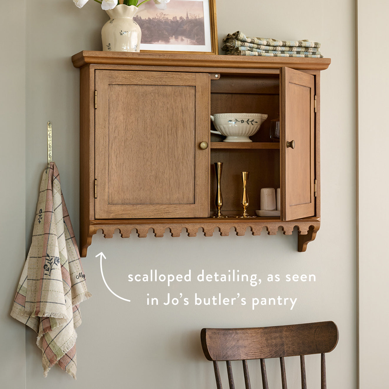 Wooden wall-mounted cabinet with scalloped detailing, featuring a plaid towel and chair in a room setting.