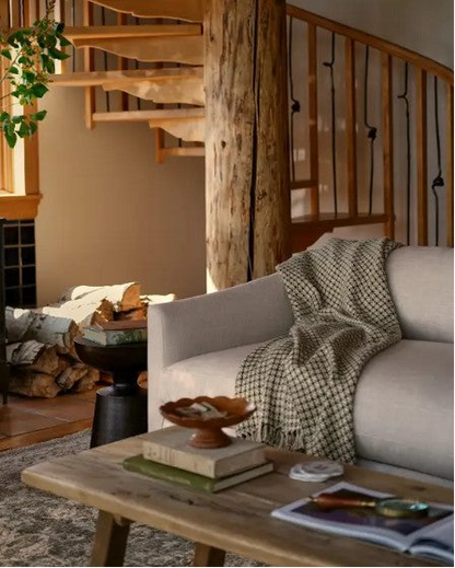 A cozy living room scene with a light beige sofa draped with a black-and-white checkered throw blanket, a rustic wooden coffee table, a black wood-burning stove with a visible fire, stacked firewood, and a spiral staircase in the background.