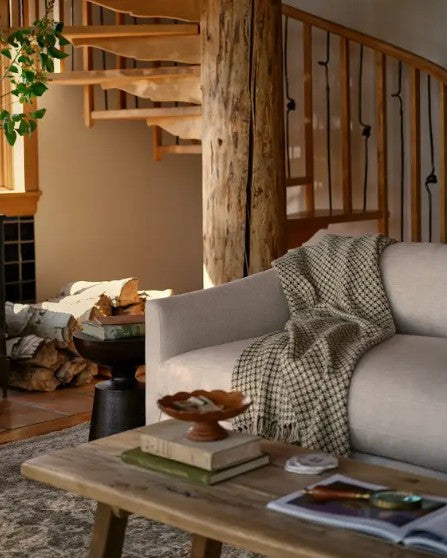 A cozy living room scene with a light beige sofa draped with a black-and-white checkered throw blanket, a rustic wooden coffee table, a black wood-burning stove with a visible fire, stacked firewood, and a spiral staircase in the background.