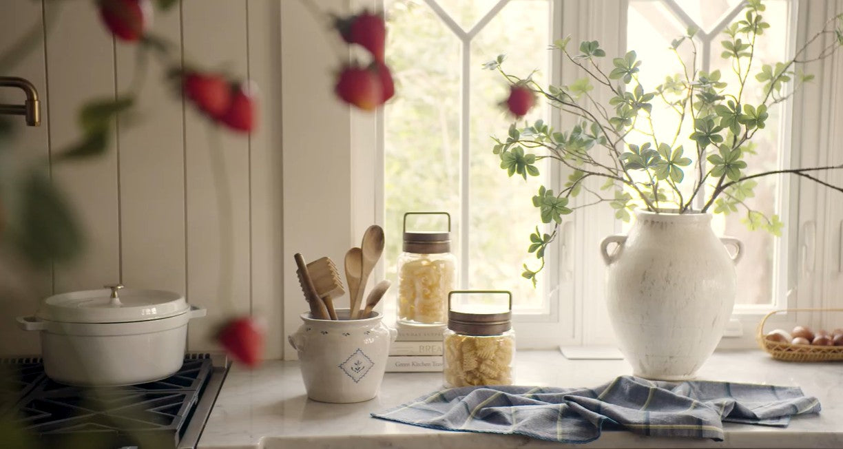 Kitchen counter with jars, utensils, and a vase with flowers near a window.
