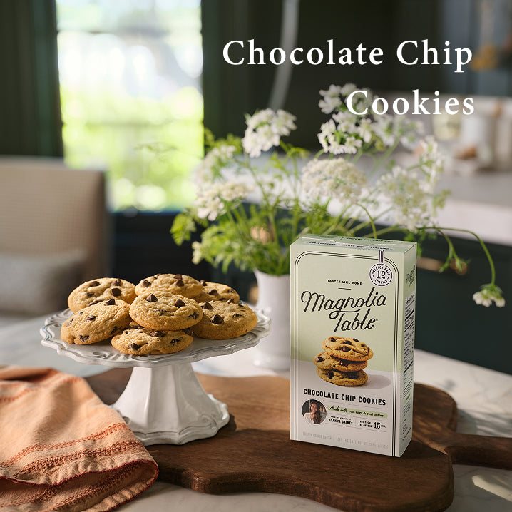 Box of Magnolia Table chocolate chip cookies on a table with cookies and flowers in the background.