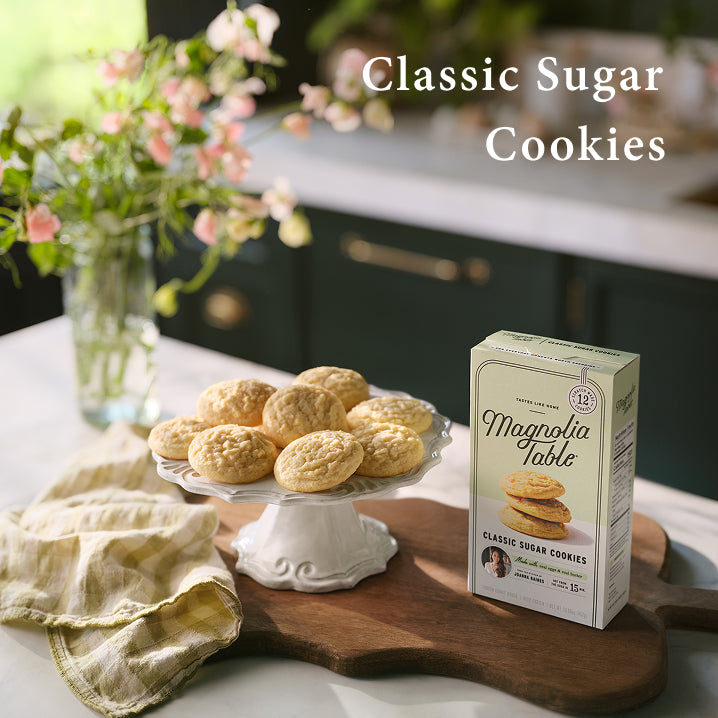 Box of Magnolia Table Classic Sugar Cookies on a kitchen counter with cookies and flowers in the background.