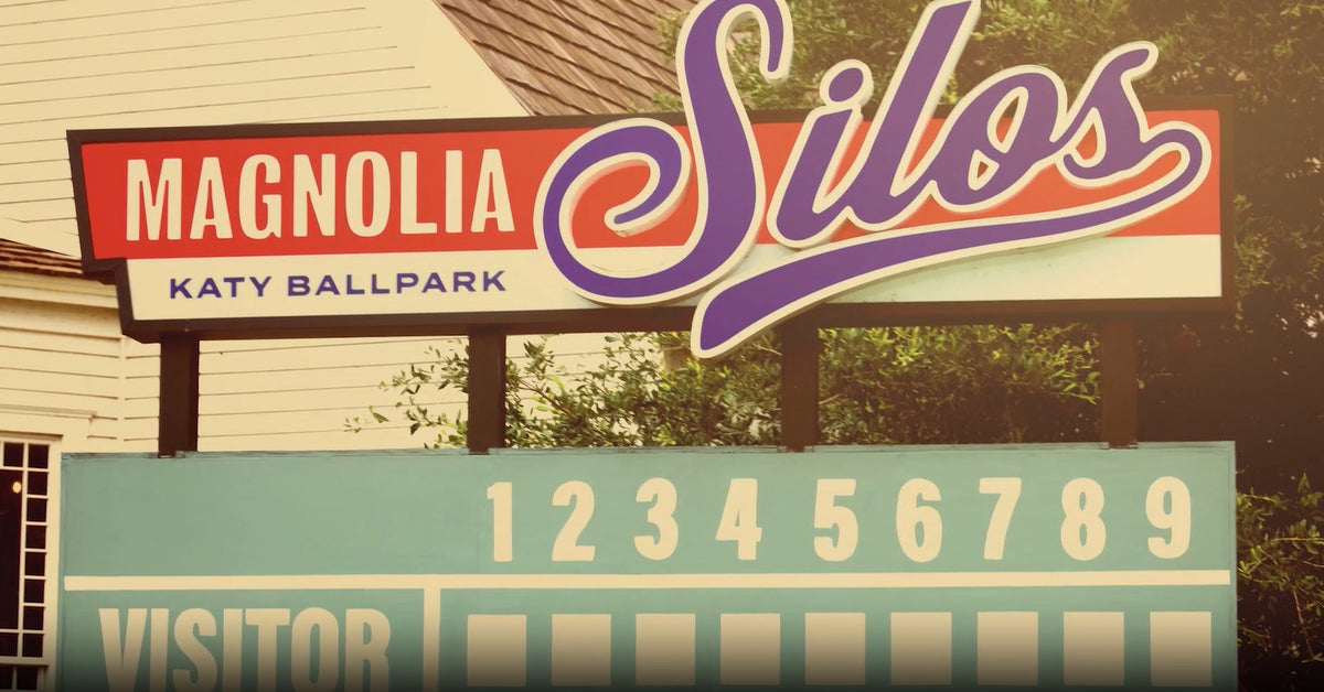 A vintage-style outdoor sign for Magnolia Silos at Katy Ballpark, with a red, white, and purple sign on top and a teal visitor number board below, set against a house and trees.