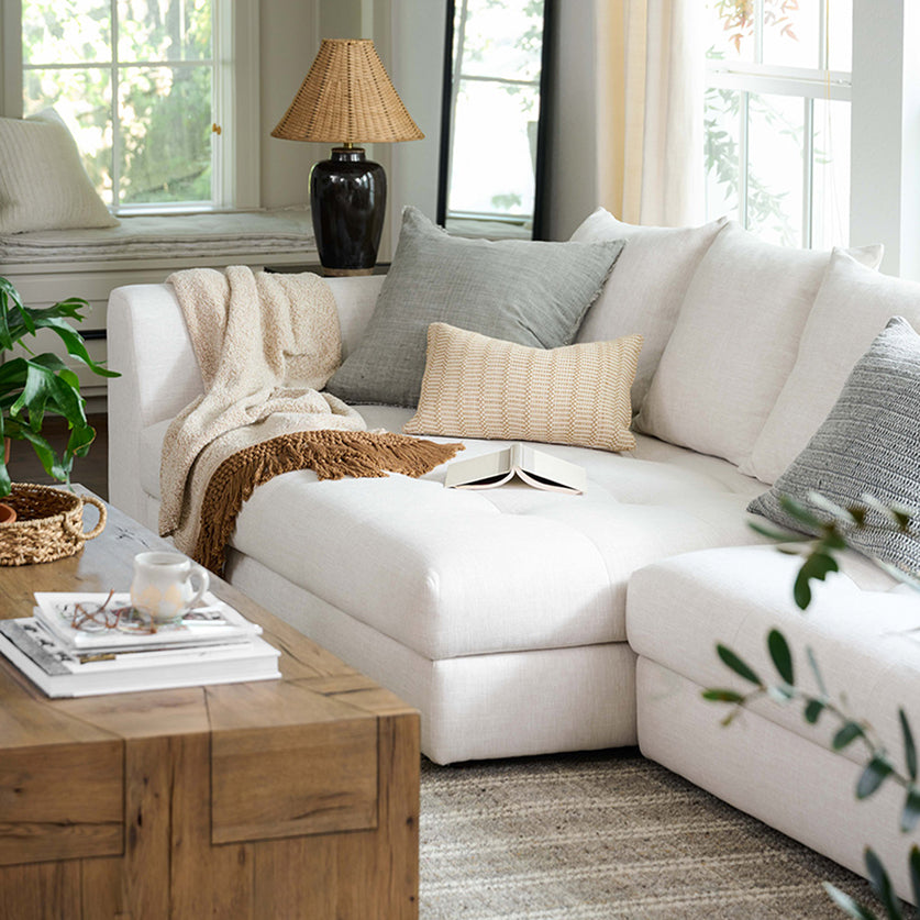 A white upholstered sectional sofa in a bright, modern living room, styled with neutral throw pillows, a cream knit blanket, a woven rug, and a wooden coffee table.