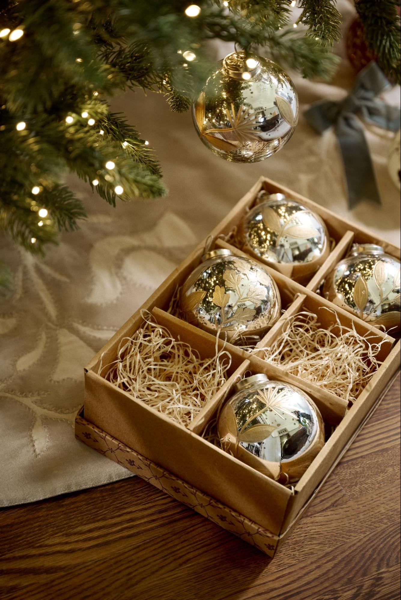 Silver bulb ornaments are packed in a brown box next to a Christmas tree.