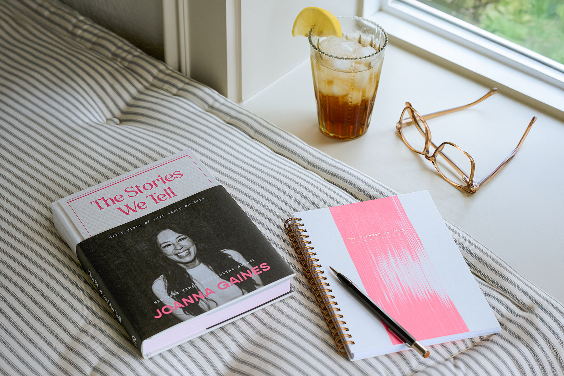 The Stories We Tell by Joanna Gaines sits on a cushion with a notebook, cup of iced tea, and pair of glasses.