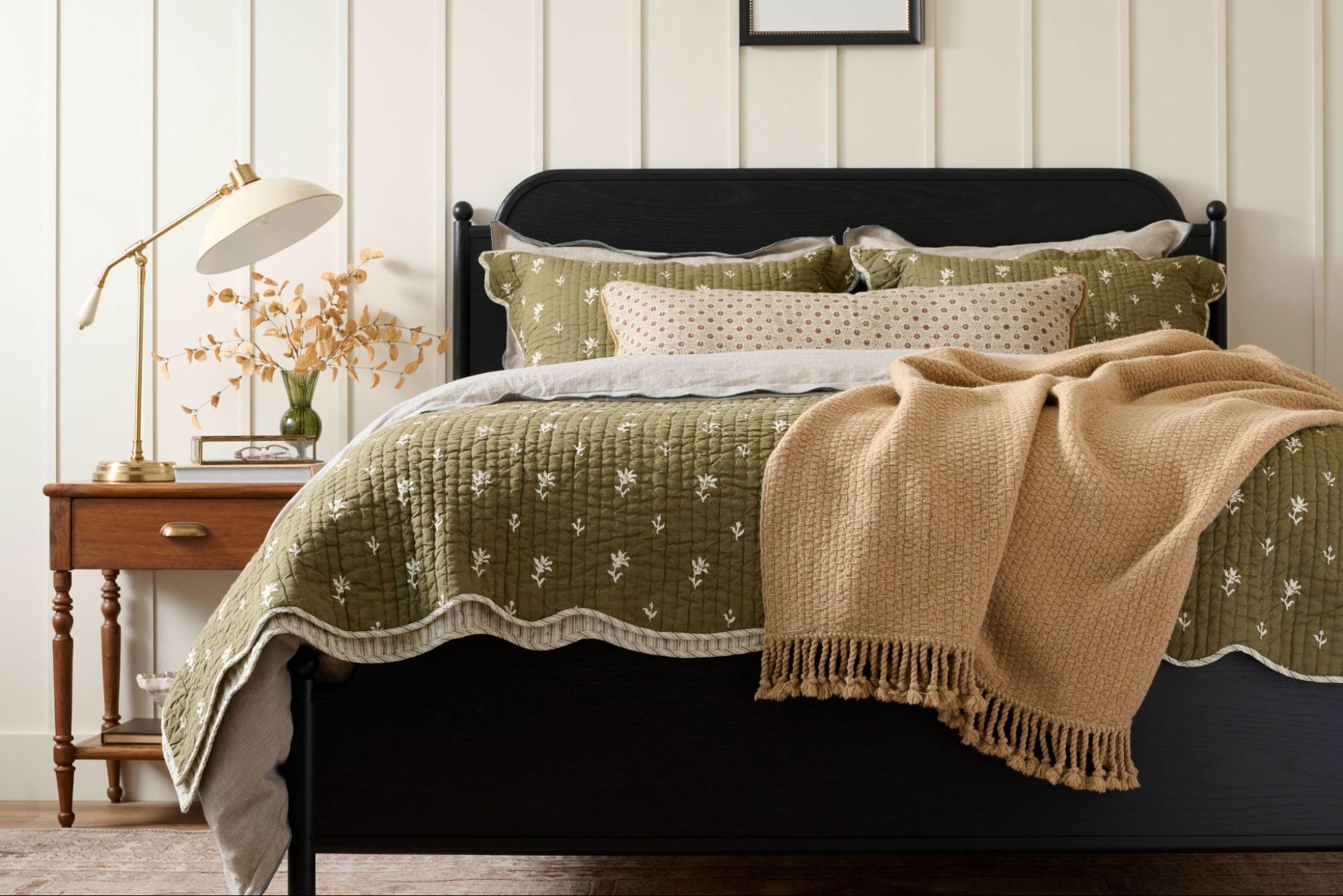 Black bed frame with green quilt next to a wooden nightstand and lamp.
