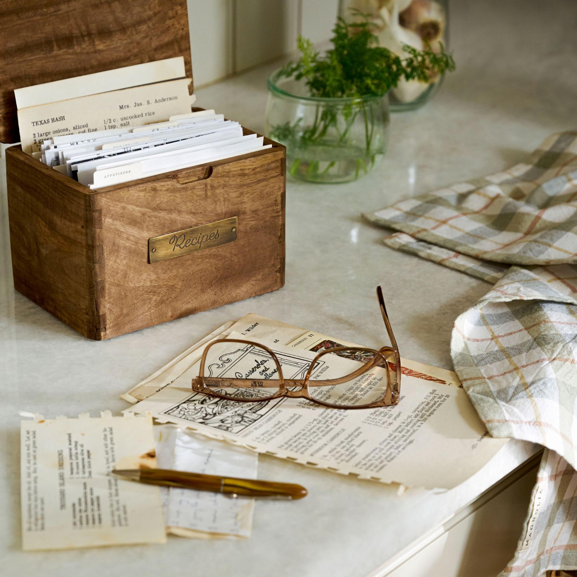 Wooden recipe box is open to reveal recipe cards. A pair of glasses and pen rest on top of displayed recipe cards.