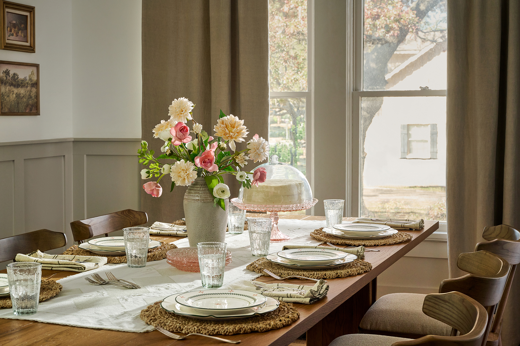 A white cake and a bouquet of pink and white flowers sit on a table set with Magnolia dishes and glasses.