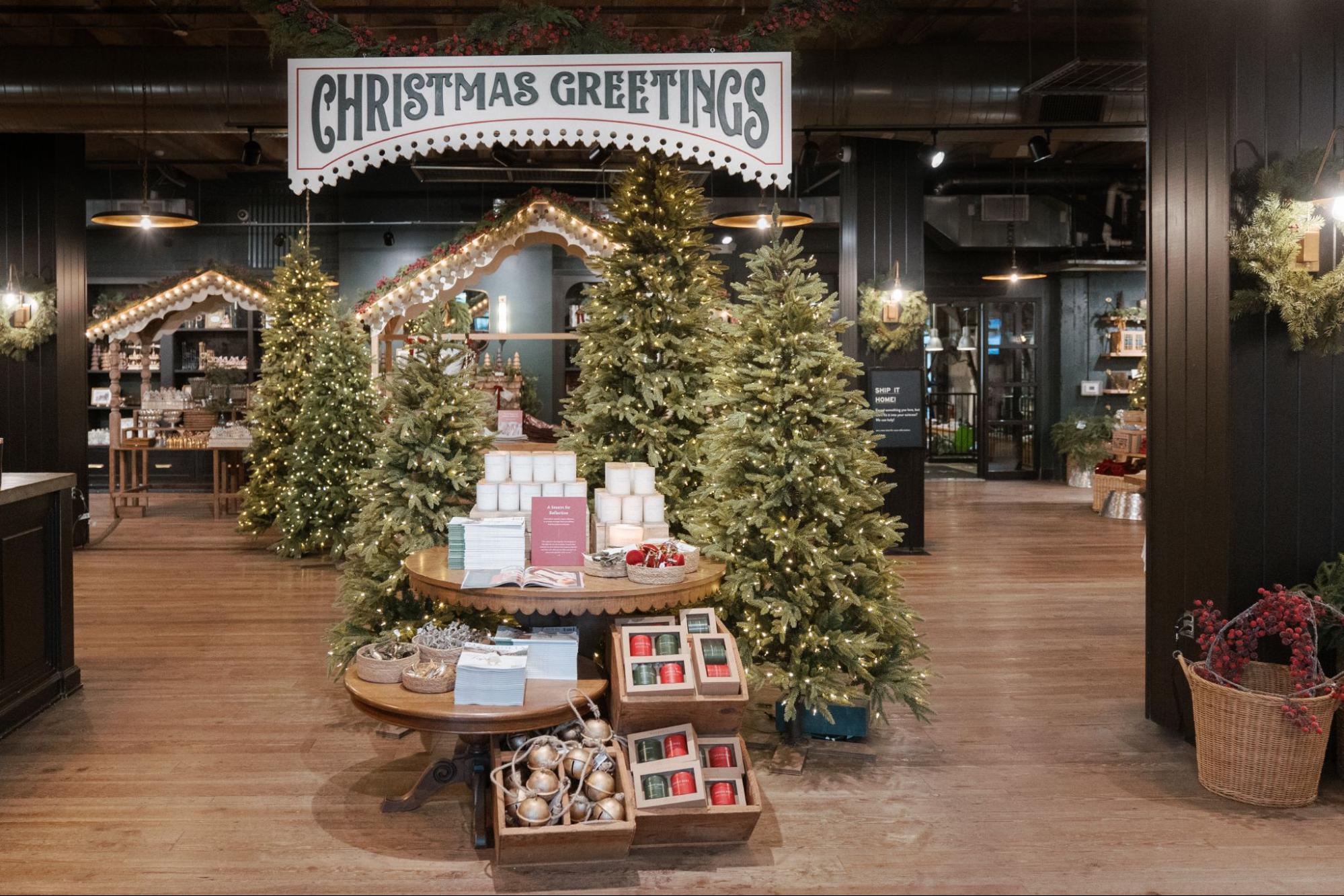 Faux evergreen trees surround a table with Magnolia merchandise—topped by a sign that says "Christmas Greetings."