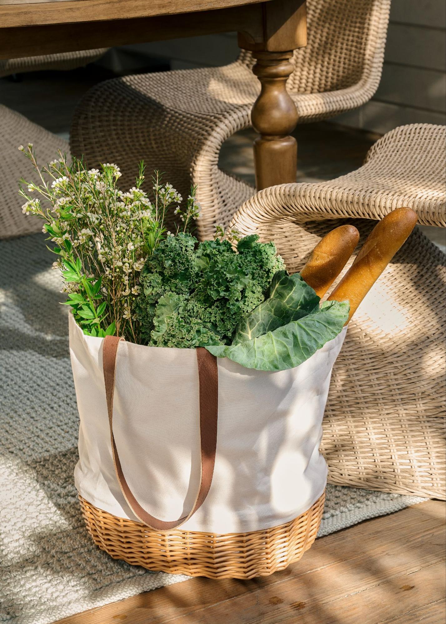 A white and wicker tote bag is filled with leafy greens, flowers, and bread—sitting in the sunlight.