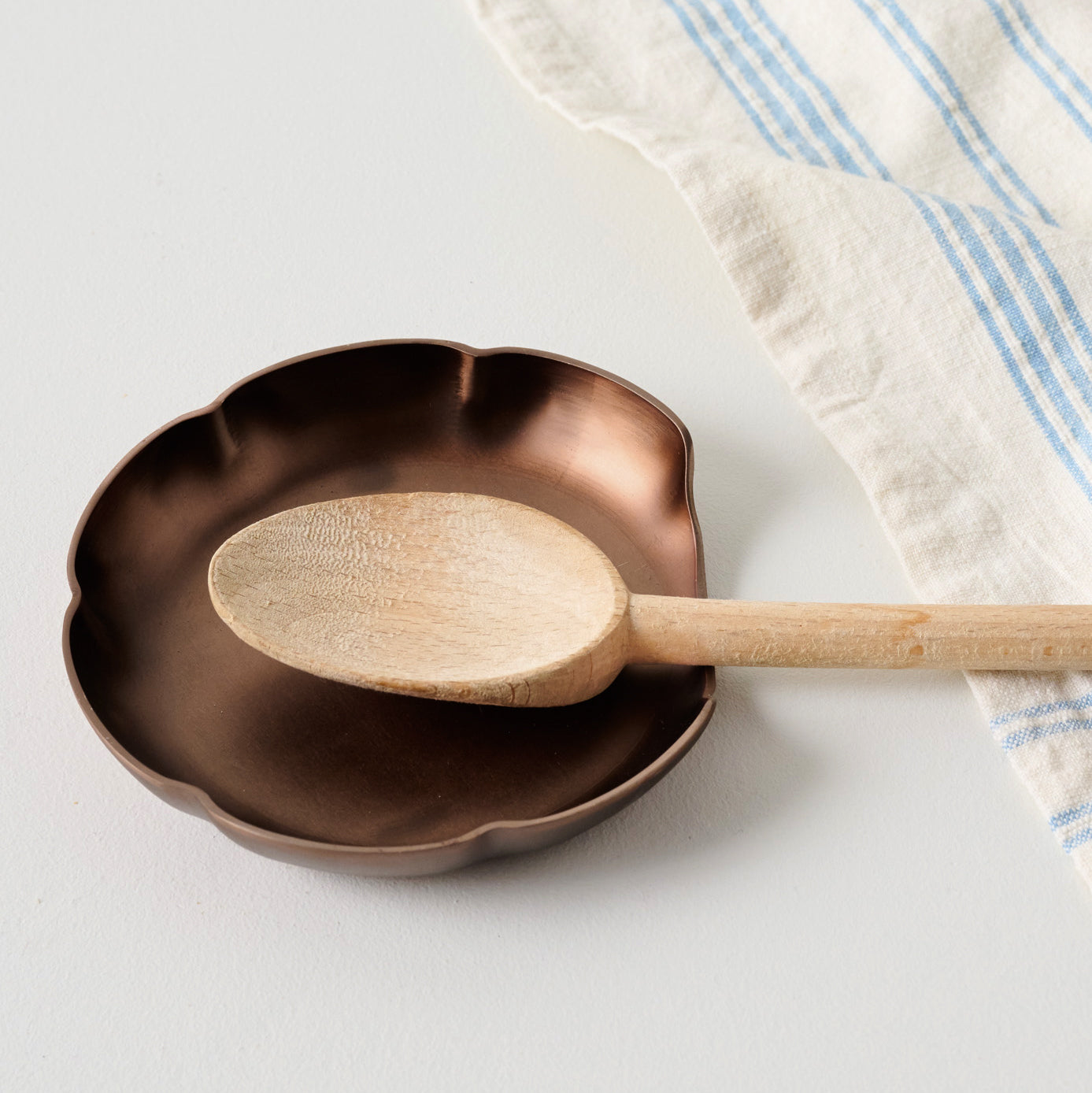 Copper Scalloped Spoon Rest shown with wooden spoon and tea towel